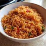 Close-up of a white bowl filled with vibrant orange spanish rice mixed with peas and red peppers.
