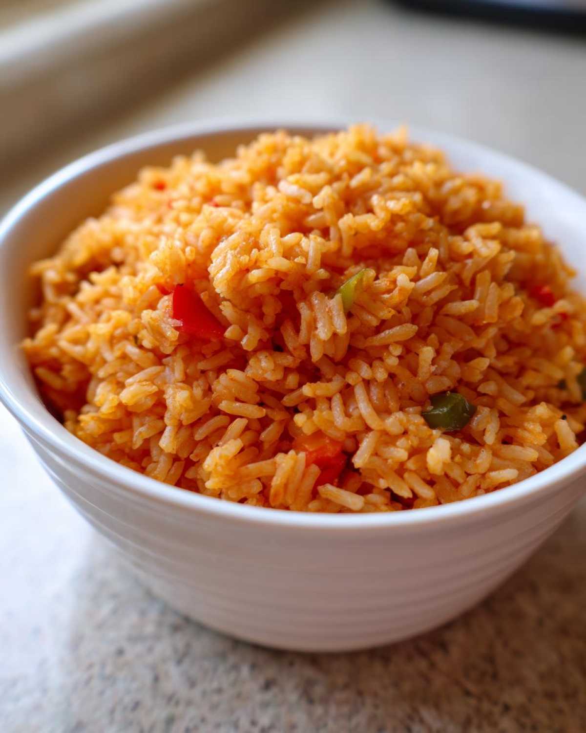 Close-up of fluffy, orange-colored spanish rice mixed with small pieces of red and green peppers in a white bowl.