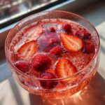 Close-up of a glass filled with sparkling pink champagne punch garnished with fresh raspberries and sliced strawberries.