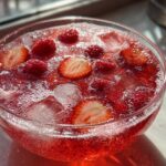 A close-up of a bright red, fizzy champagne punch garnished with fresh raspberries and sliced strawberries over ice.
