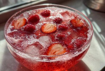 A close-up of a bright red, fizzy champagne punch garnished with fresh raspberries and sliced strawberries over ice.