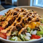 A close-up of a fresh cheeseburger bowl featuring lettuce, tomatoes, seasoned ground beef, melted cheese, and a drizzle of sauce.