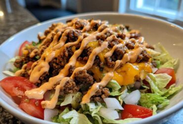 A close-up of a fresh cheeseburger bowl featuring lettuce, tomatoes, seasoned ground beef, melted cheese, and a drizzle of sauce.