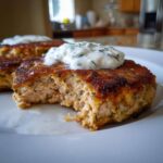 A close-up view of a golden-brown salmon cake, cut open to show the flaky interior, topped with creamy dill sauce.