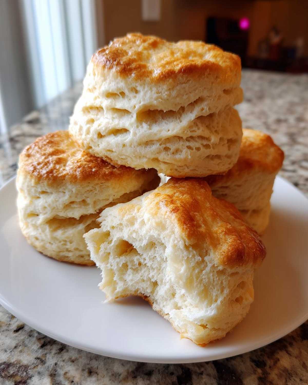 A stack of four golden-brown, tall buttermilk biscuits, one broken open to show the fluffy interior.