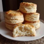 A stack of tall, golden-brown buttermilk biscuits with one biscuit broken open to show the fluffy interior.