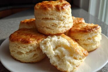 A stack of tall, golden-brown buttermilk biscuits on a white plate, one broken open to show fluffy interior.