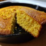A close-up shot showing a fluffy slice of golden buttermilk cornbread being lifted from the rest of the cake in a black cast-iron skillet.