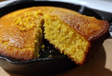 A close-up shot showing a fluffy slice of golden buttermilk cornbread being lifted from the rest of the cake in a black cast-iron skillet.