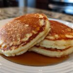 A close-up of a stack of fluffy greek yogurt pancakes drizzled with golden syrup on a white plate.