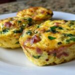 Close-up of three fluffy muffin tin egg bites with visible ham and green spinach on a white plate.