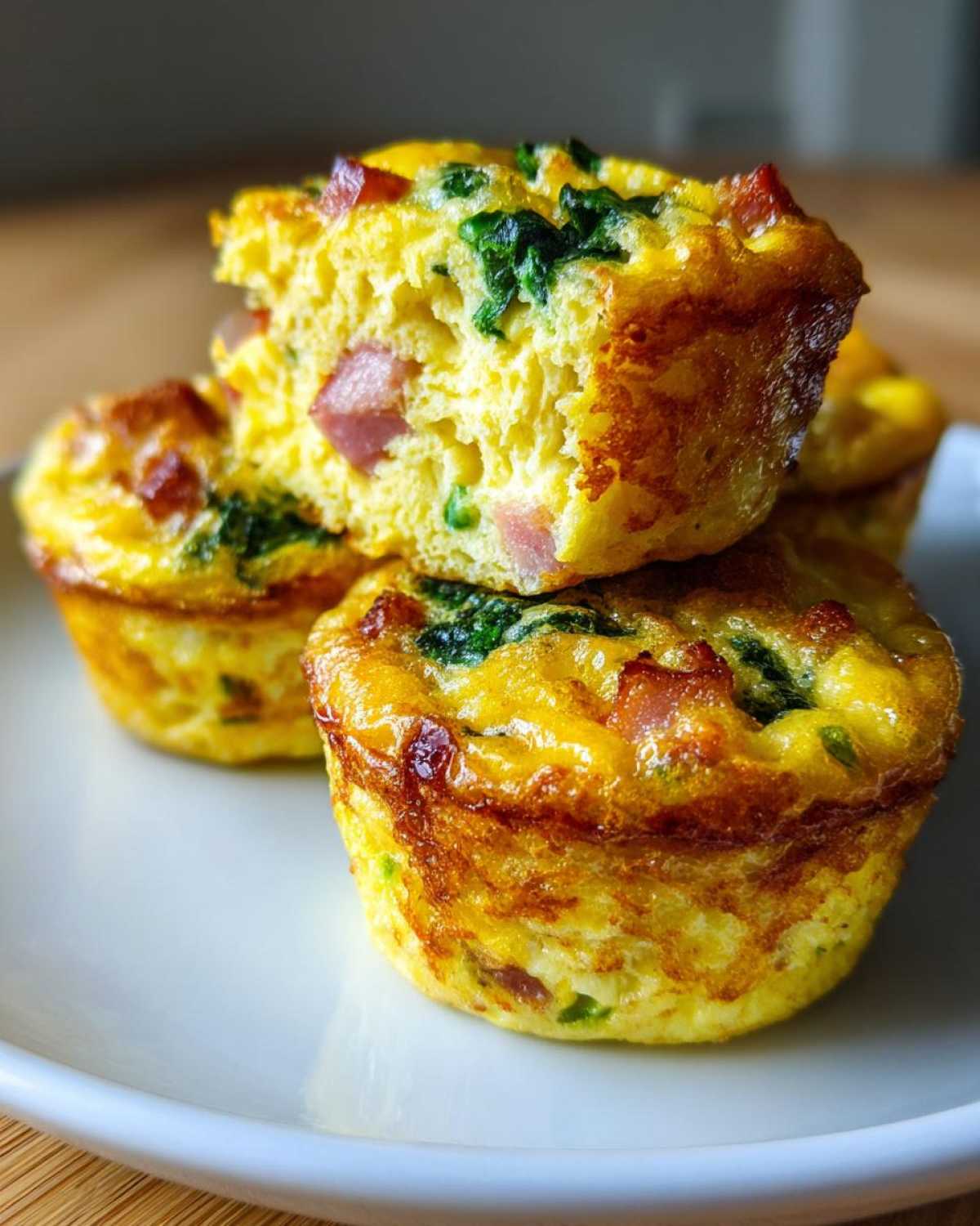Close-up of fluffy muffin tin egg bites stacked on a white plate, showing ham and spinach inside the baked egg mixture.