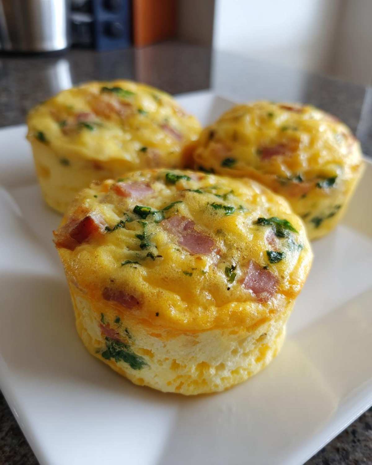 Close-up of three fluffy muffin tin egg bites containing ham and green herbs served on a white square plate.