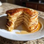 A tall stack of fluffy sourdough pancakes with a slice removed, topped with melting butter and drizzled with syrup.