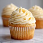 Close-up of a perfectly baked vanilla cupcake topped with a swirl of creamy vanilla frosting, with more vanilla cupcakes blurred in the background.