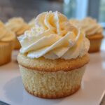 A close-up of one perfectly baked vanilla cupcake topped with a swirl of pale yellow buttercream frosting, with more vanilla cupcakes blurred in the background.