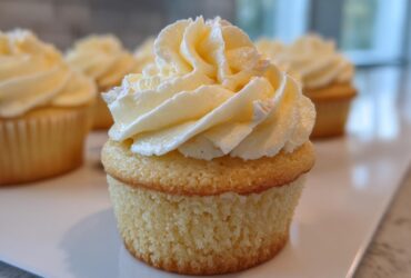 A close-up of one perfectly baked vanilla cupcake topped with a swirl of pale yellow buttercream frosting, with more vanilla cupcakes blurred in the background.