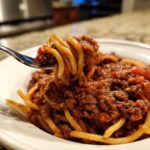 Close-up of a fork lifting spaghetti coated heavily in rich bolognese sauce from a white bowl.