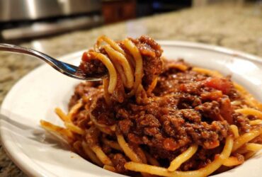 Close-up of a fork lifting spaghetti coated heavily in rich bolognese sauce from a white bowl.