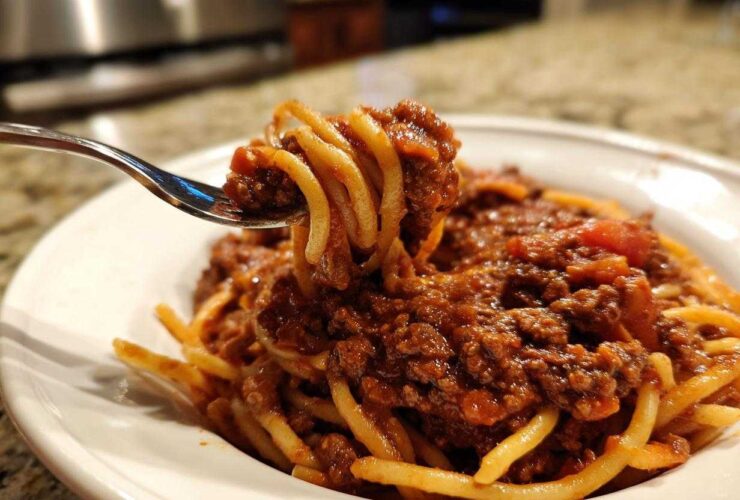 Close-up of a fork lifting spaghetti coated heavily in rich bolognese sauce from a white bowl.