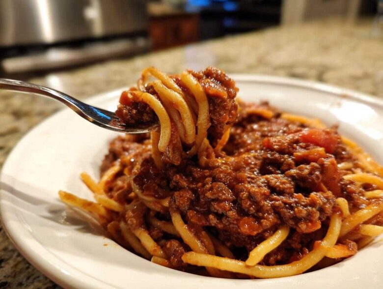 Close-up of a fork lifting spaghetti coated heavily in rich bolognese sauce from a white bowl.