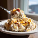 A fork lifts a generous scoop of creamy baked potato salad topped with shredded cheddar and bacon bits.