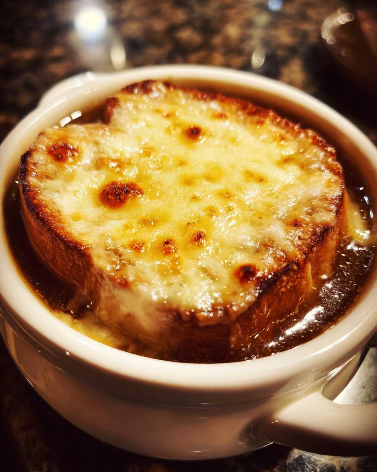 Close-up of a bowl of rich french onion soup recipe topped with a thick, melted, and browned cheese crust over bread.