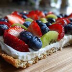 Close-up side view of a delicious fruit pizza sugar cookie topped with cream cheese frosting, strawberries, blueberries, and kiwi.