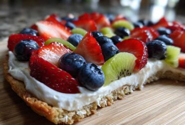 Close-up side view of a delicious fruit pizza sugar cookie topped with cream cheese frosting, strawberries, blueberries, and kiwi.