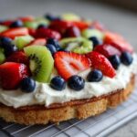 A close-up view of a fruit pizza sugar cookie base topped with cream cheese frosting and fresh strawberries, blueberries, and kiwi slices.