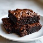 Two rich, dark, fudgy black bean brownies stacked on a white plate, topped with chocolate chips.