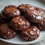 Close-up of several fudgy brownie cookies piled on a white plate, showing their signature crackled, shiny tops.