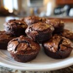 A close-up of several fudgy brownie cookies stacked on a white plate, showing their cracked, shiny tops.
