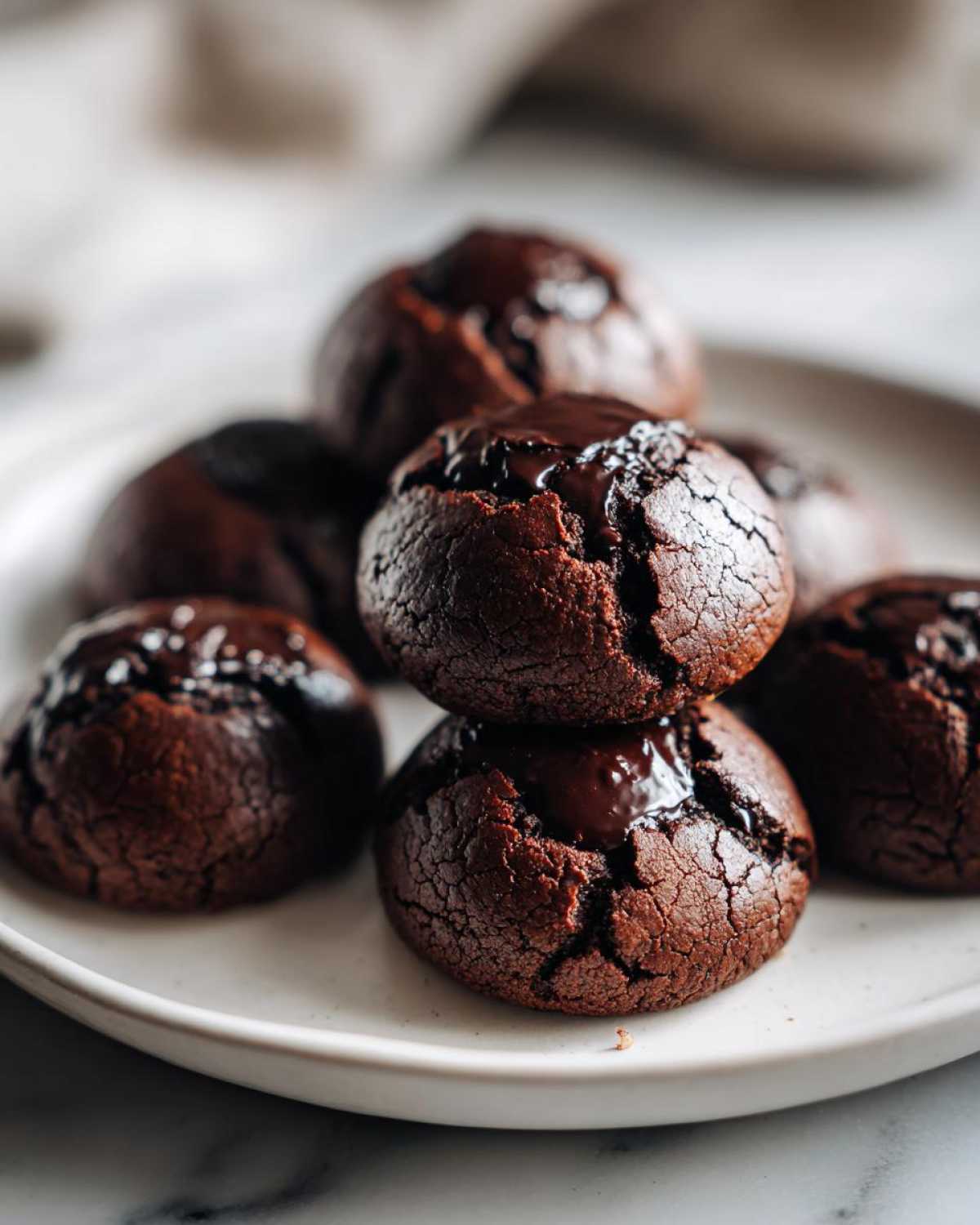 Close-up of several fudgy brownie cookies stacked on a white plate, featuring cracked tops and melted chocolate centers.