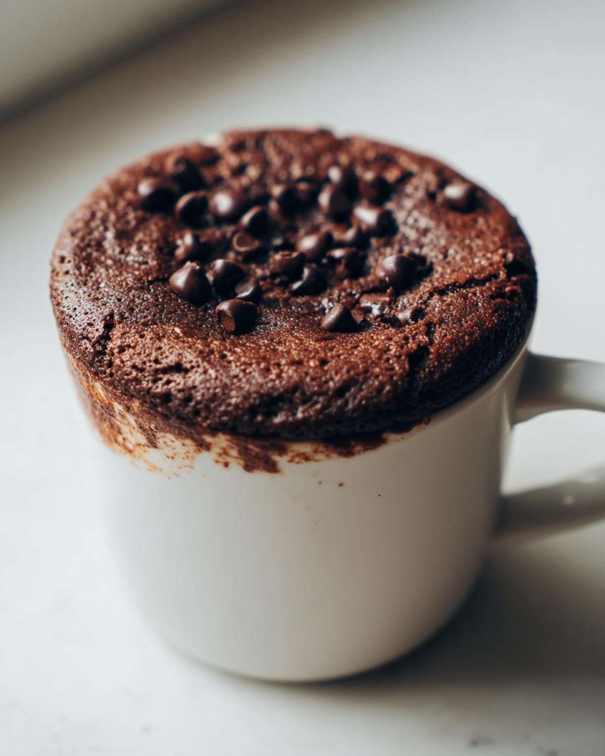 Close-up of a rich, fudgy chocolate mug cake overflowing slightly from a white mug, topped with chocolate chips.