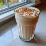 A tall, textured glass filled with creamy, frothy coquito topped generously with ground cinnamon.