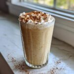 A tall glass filled with creamy, spiced coquito topped with whipped cream and a dusting of cinnamon.