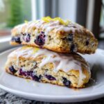 Two freshly baked blueberry scones stacked, drizzled with white glaze and topped with lemon zest.