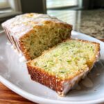 A loaf of moist lemon zucchini bread drizzled with white glaze, with one slice cut and resting beside it.