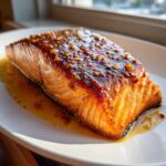 Close-up of a perfectly cooked, glazed salmon recipe fillet resting in its sauce on a white plate.