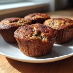 Four freshly baked banana protein muffins with golden-brown tops resting on a white plate near a window.