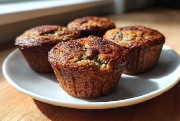 Four freshly baked banana protein muffins with golden-brown tops resting on a white plate near a window.