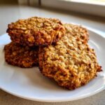 A stack of three golden brown, textured breakfast cookie rounds made with oats, served on a white plate.
