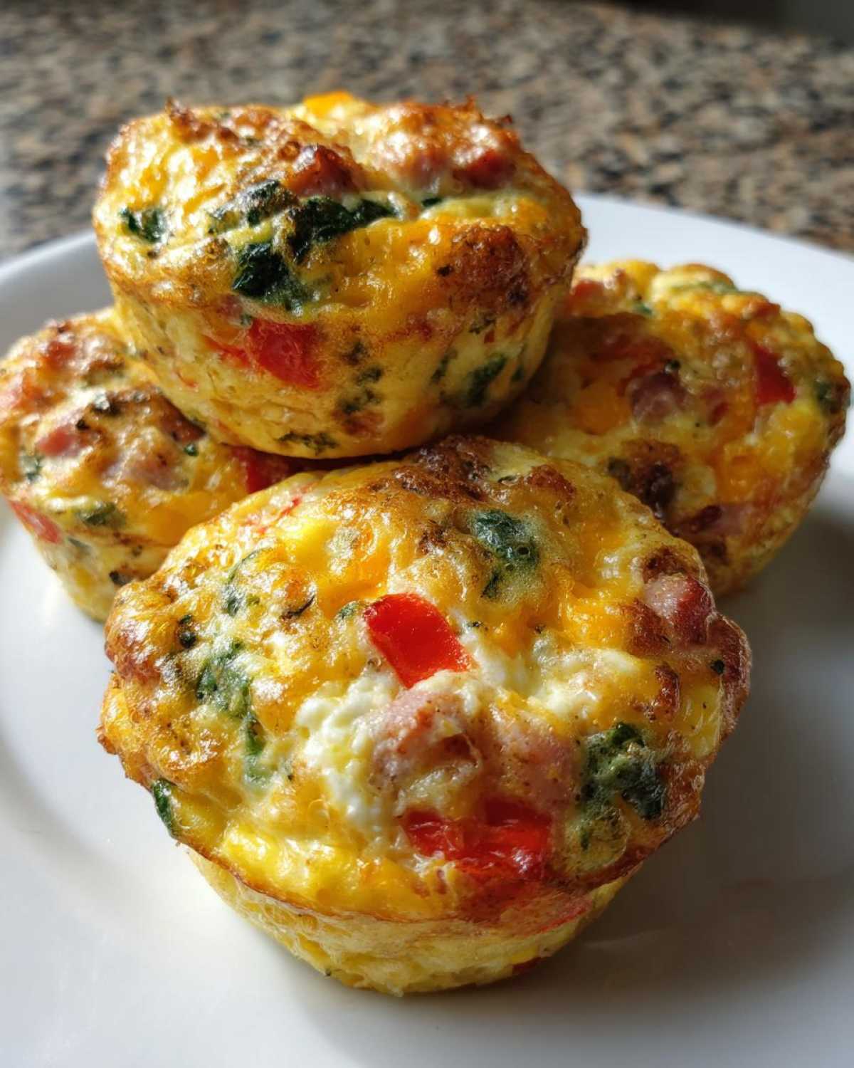 A close-up of four freshly baked Egg Muffin Cups filled with ham, red peppers, and spinach on a white plate.