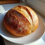 A perfectly baked, round loaf of golden brown French bread with a scored top resting on a white plate.