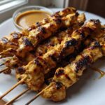 A close-up of several perfectly grilled chicken satay skewers served on a white plate next to a small bowl of peanut sauce.