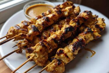 A close-up of several perfectly grilled chicken satay skewers served on a white plate next to a small bowl of peanut sauce.