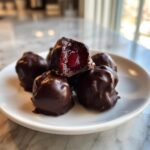 A close-up of several homemade chocolate covered cherries piled on a white plate, one is bitten open revealing the bright red cherry inside.