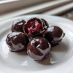 Close-up of five homemade chocolate covered cherries on a white plate, one is broken open showing the red cherry inside.
