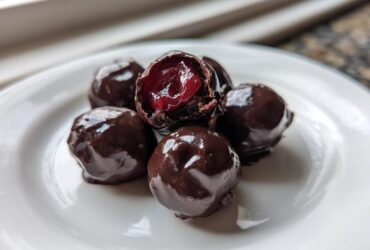 Close-up of five homemade chocolate covered cherries on a white plate, one is broken open showing the red cherry inside.